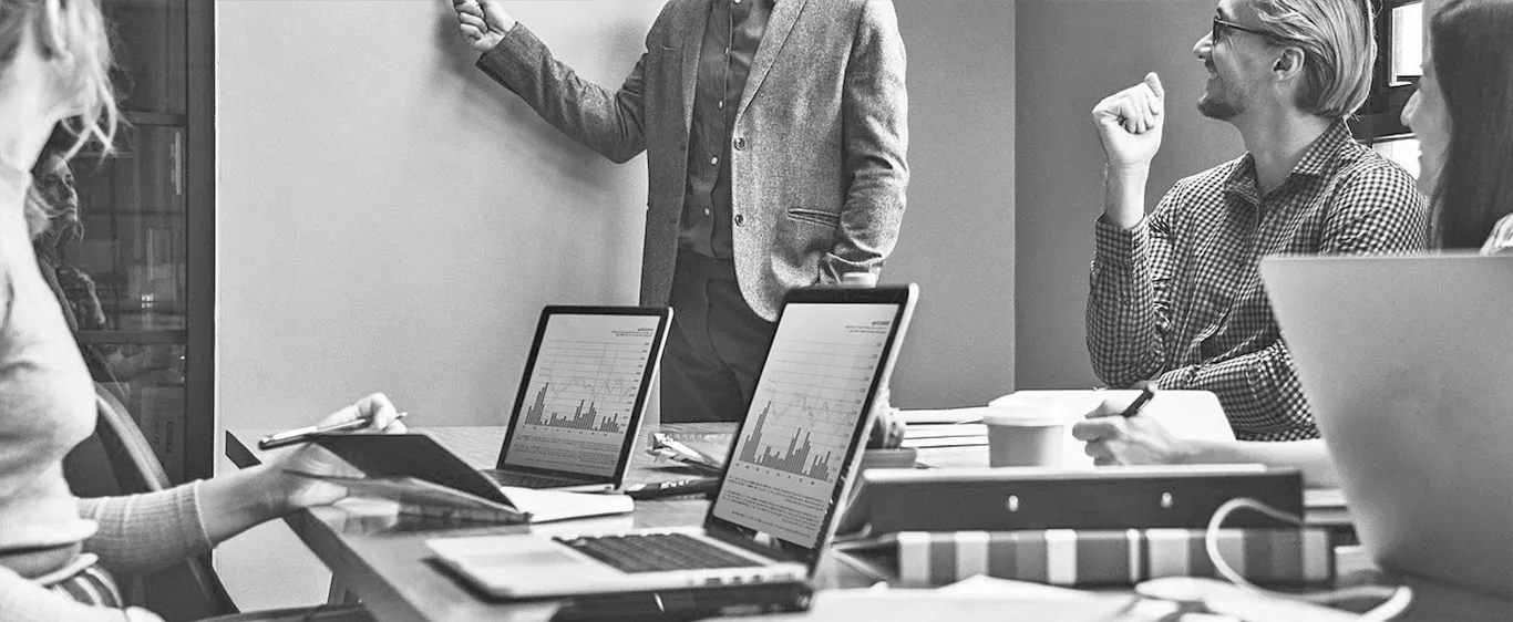 Multiple people meeting int a conference room white boarding ideas and looking at analytics graphs on their laptops.